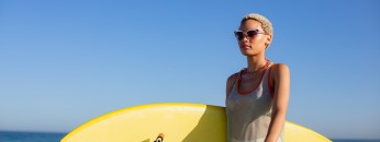 Front view of African american woman with surfboard sitting on bicycle at beach in the sunshine