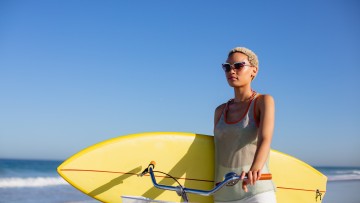 Front view of African american woman with surfboard sitting on bicycle at beach in the sunshine