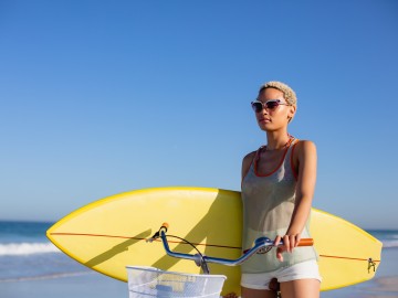 Front view of African american woman with surfboard sitting on bicycle at beach in the sunshine